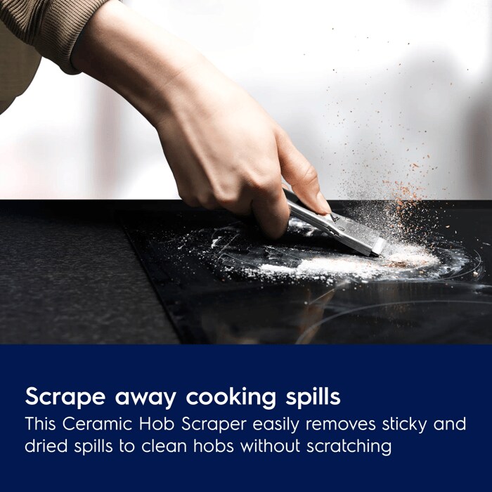 Close-up of a hand using a metal scraper to remove dried white residue and food spills from a black ceramic cooktop.